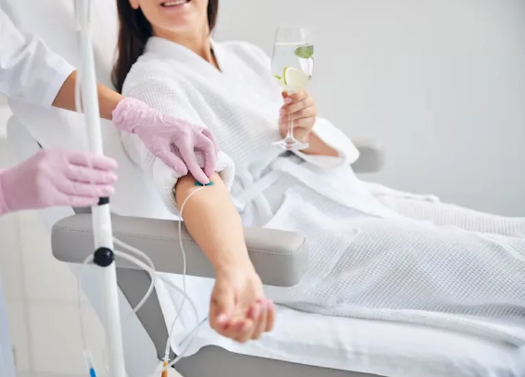 Dark-haired spa client with a glass of drink in her hand looking away during the intravenous therapy