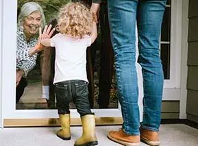 Small child visiting grandparent through glass door with parent standing nearby