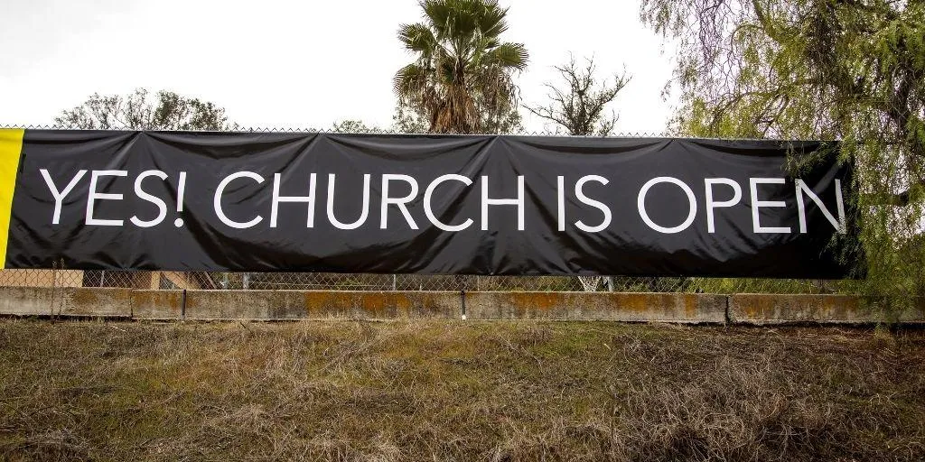 Black banner sign on fence declaring yes church is open. 