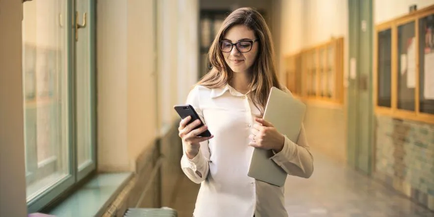 Young woman in glasses standing in hallway and smiling at phone as she uses text messaging for church.