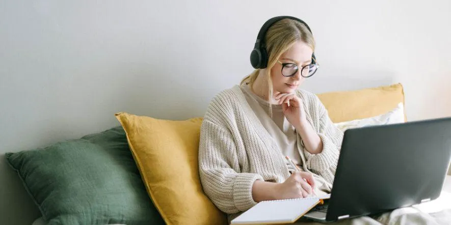 Young woman wearing headphones and jotting down notes in notebook in front of open laptop.