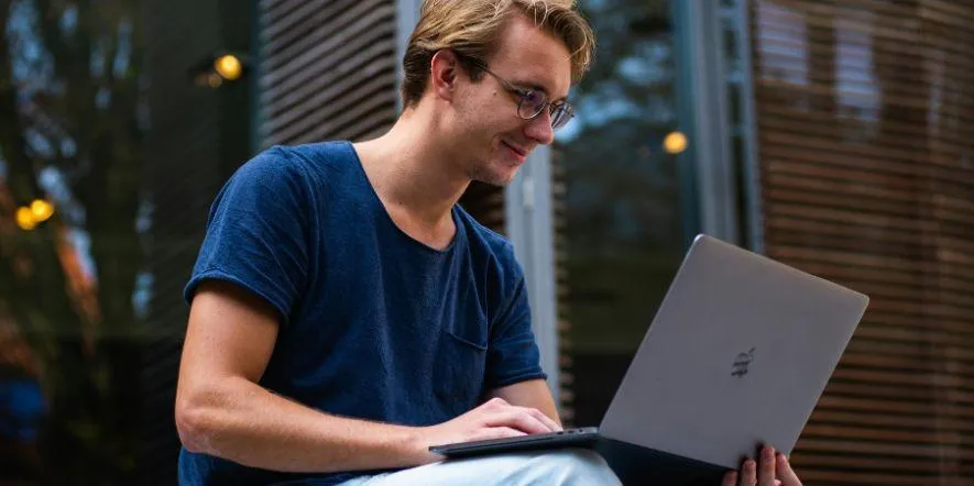 A young man in glasses sitting outside and smiling at his laptop as he engages in online prayer.
