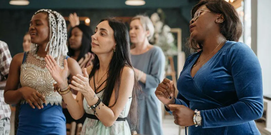 A group of women standing up from their chairs and engaging in worship during a small group gathering. 