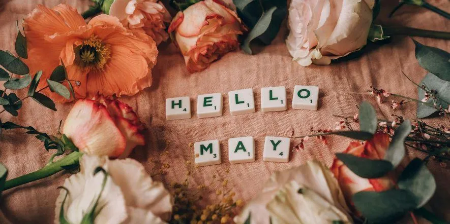The words hello May spelled out in Scrabble tiles on a wooden table covered in flowers.