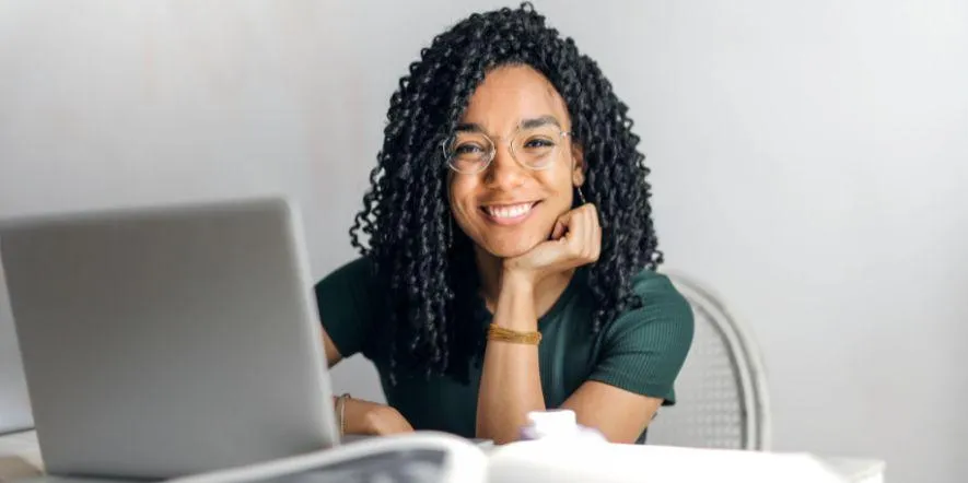 A smiling woman in glasses sitting at a desk in front of an open laptop.