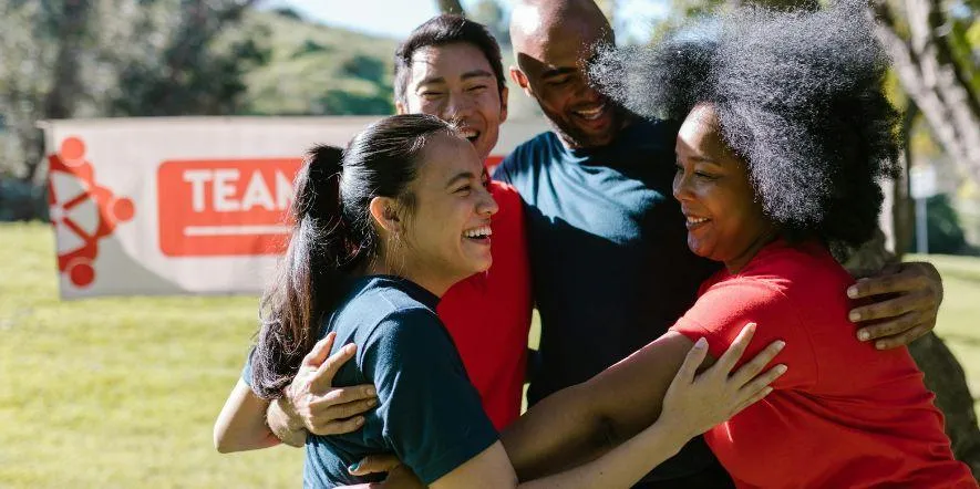 Two men and two women smiling as they join in a group hug, with a banner that says team in the background.