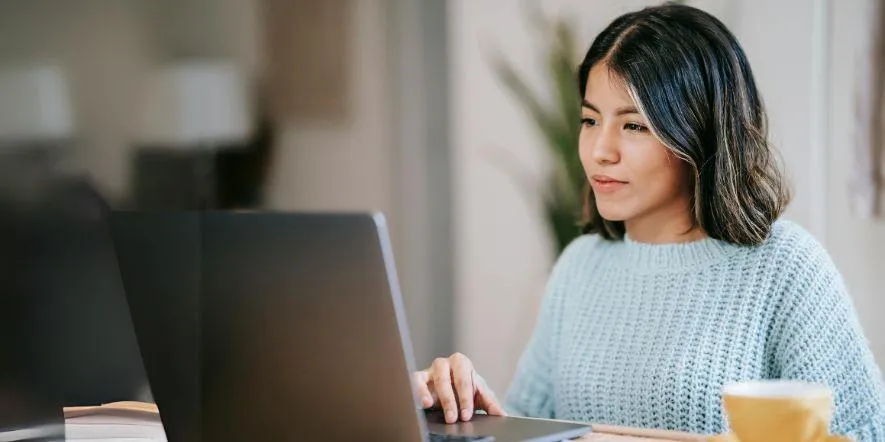 A young brunette woman in a blue sweater looking at her laptop as she engages in religious study.