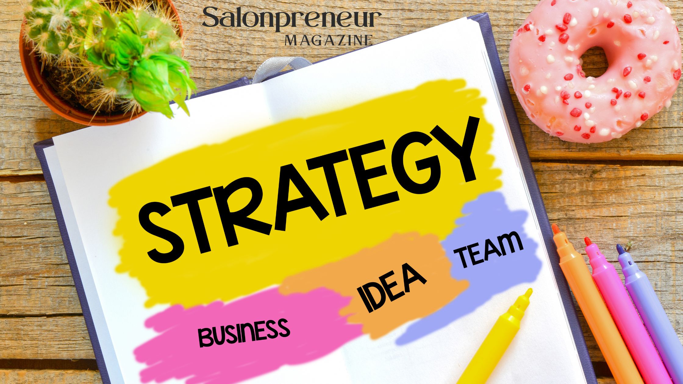 A table with a plant, pink sprinkled doughnut and a pad with colour blocks with the words strategy, business, idea and team on coloured areas, pens lay to the side. Salonpreneur Magazine logo in black centre top