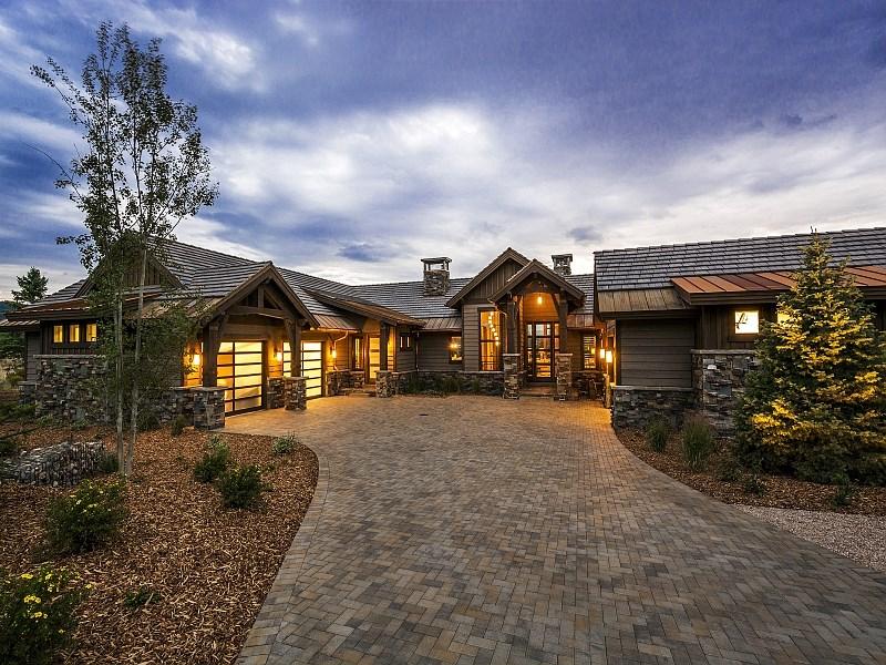 Luxury mountain-style home with warm exterior lighting and a wide paver driveway surrounded by landscaped trees and mulch beds under a dramatic evening sky.
