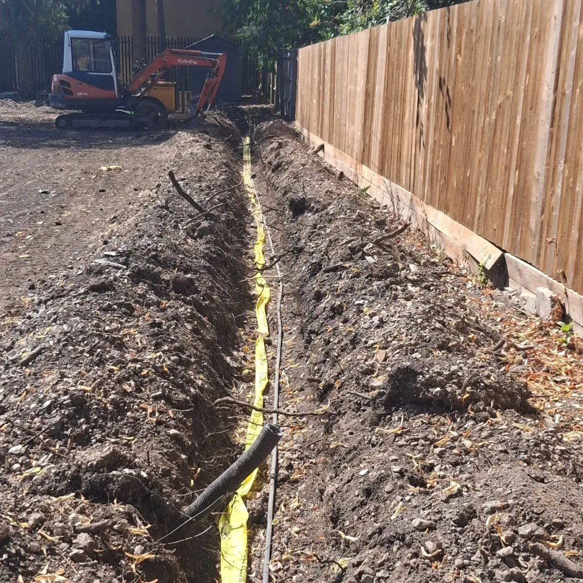Trench dug along a fence line with warning tape and service ducting, excavator in the background