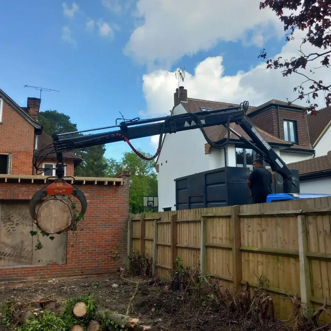 Crane grab lifting a cut tree trunk section over a garden fence into a truck