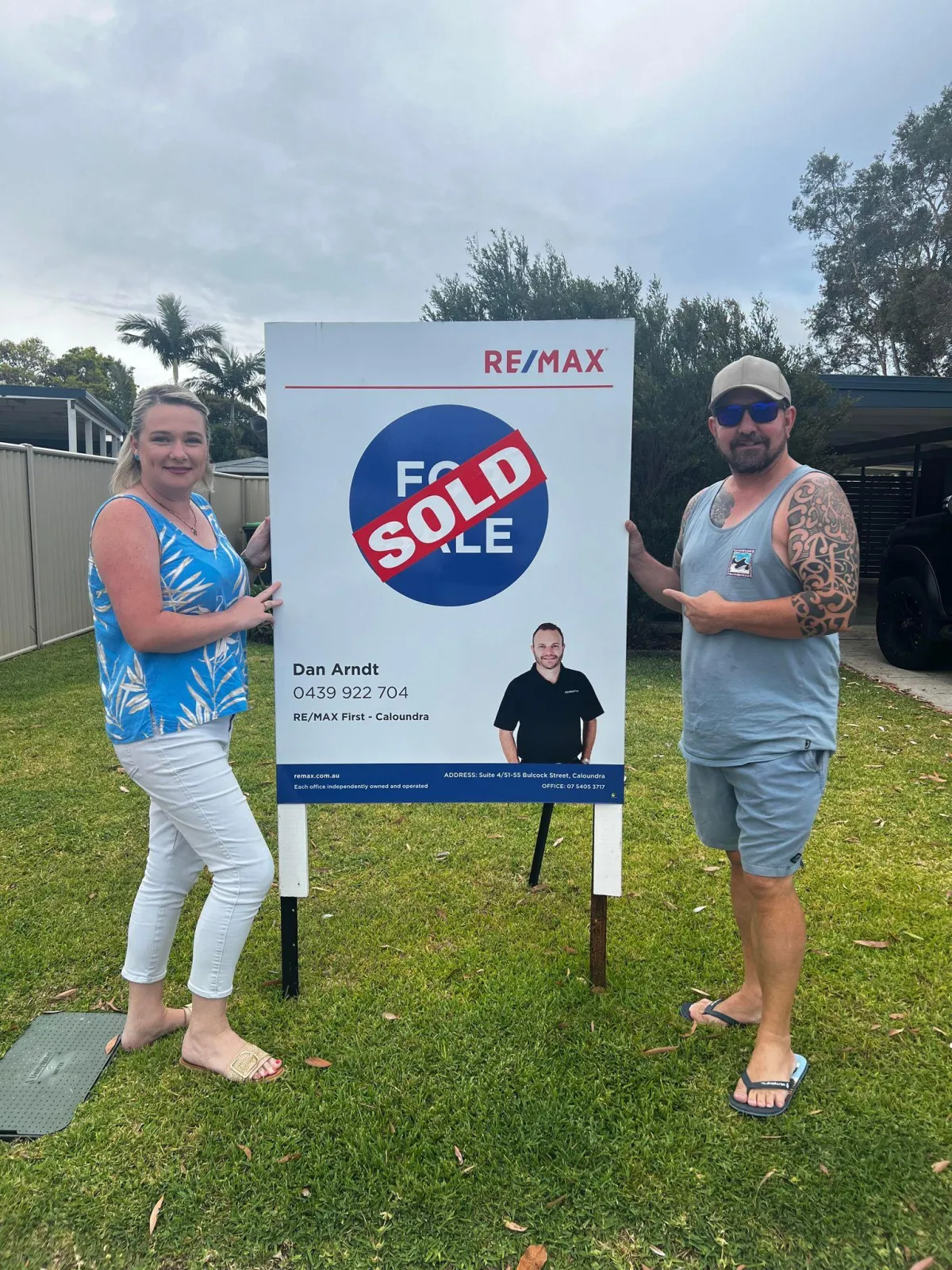 Two smiling buyers celebrate beside a sold sign in front of their new home.