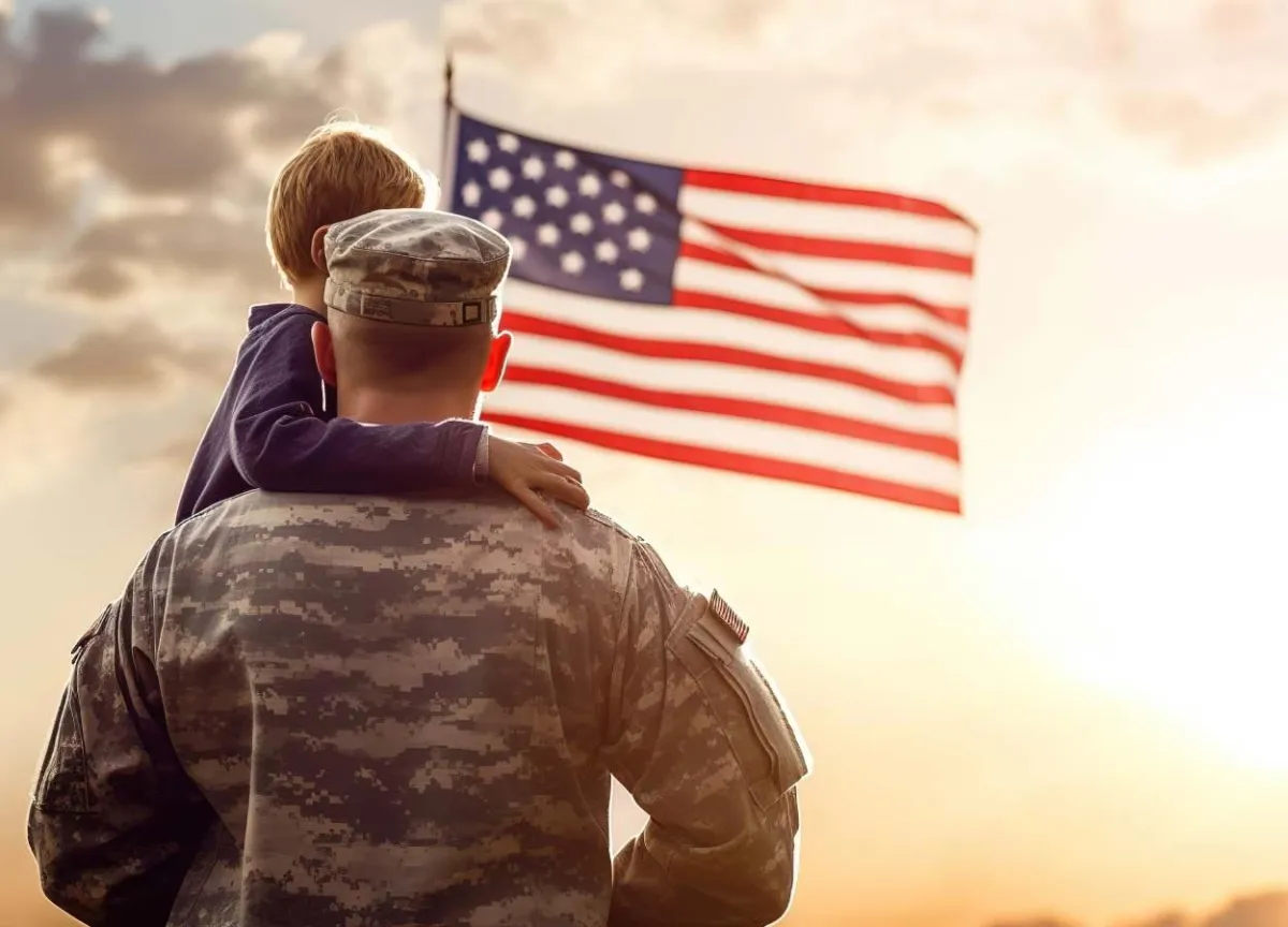 A US army soldier holding a child and looking into the sunset at an American flag