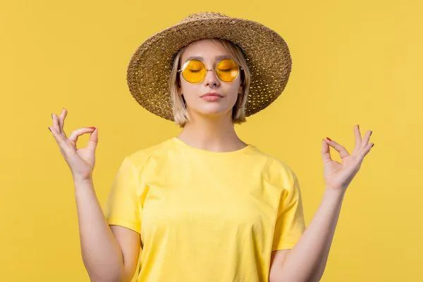 bright yellow background. A lady with yellow round sunglasses and a big hat whilst she is meditating.