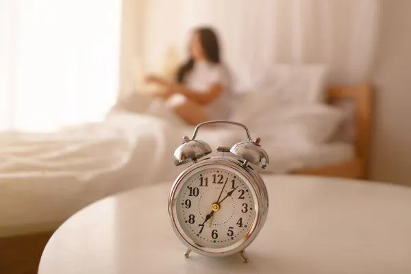 An old style alarm clock in the foreground after 7.00, with somebody sat on the bed not quite up yet.