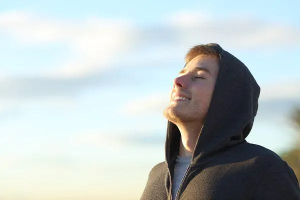 Man stood on a beach taking a deep breath. He is wearing a green hoddie with the hood up. The sky is blue with a few clouds.