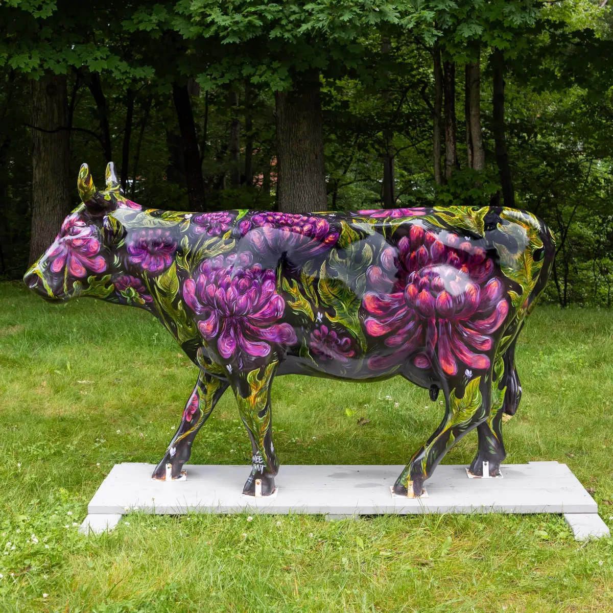A darkly painted cow sculpture featuring purple dahlia flowers and bees