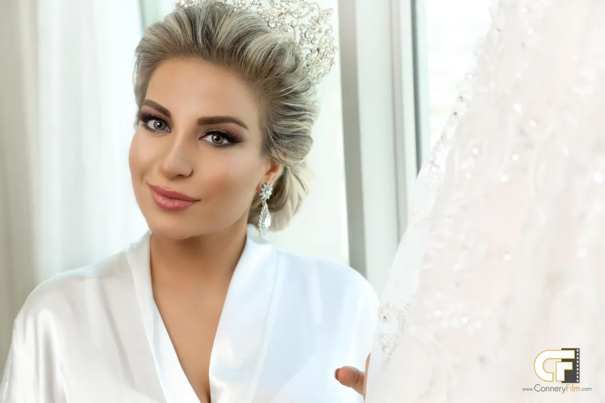 Close-up portrait of a beautiful bride with an elegant updo, tiara, and dangling earrings, wearing a white satin robe and smiling directly at the camera, with a glimpse of her wedding dress on the right.