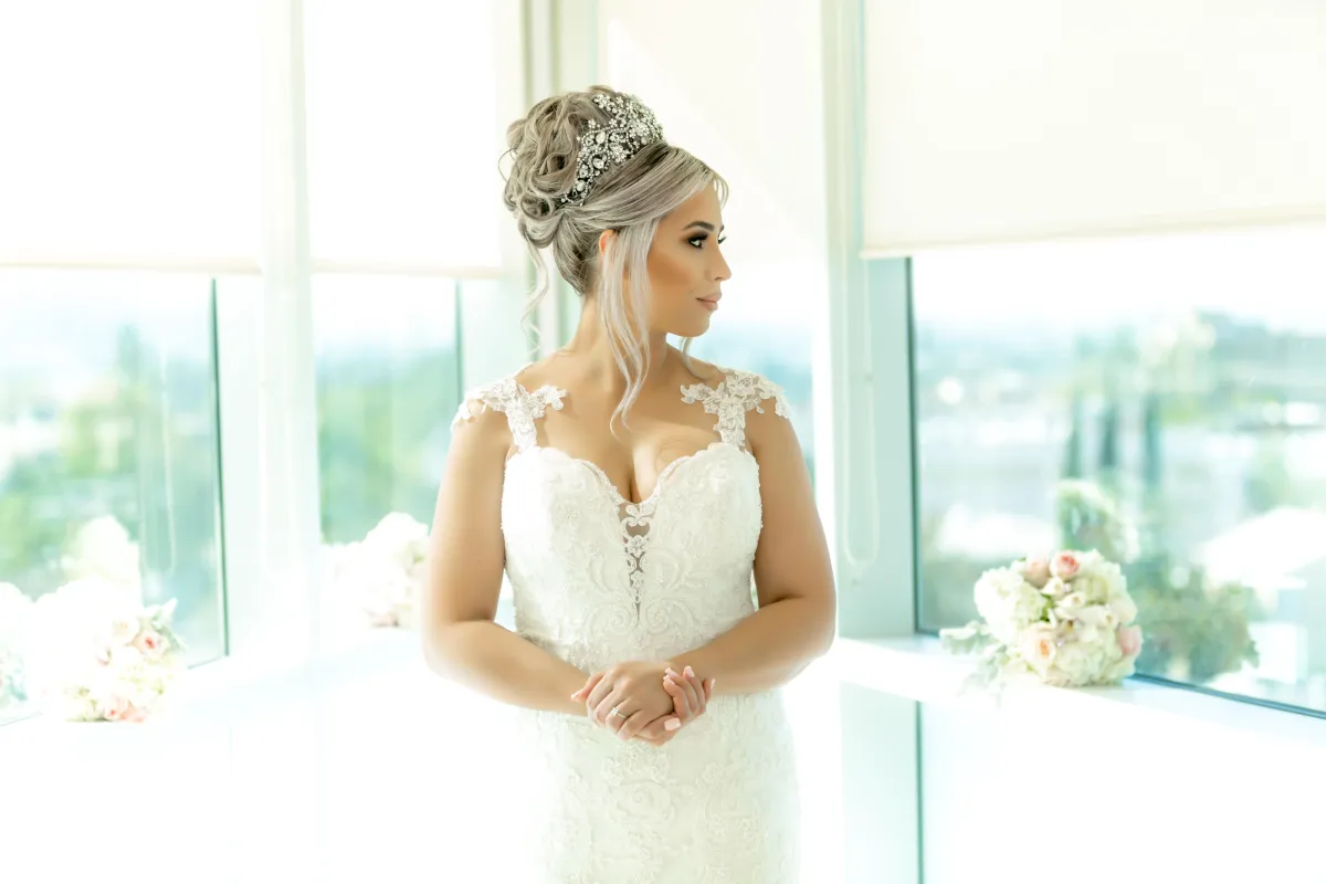 A beautiful bride in a white lace wedding dress with an elegant updo and tiara, standing by a window, looking thoughtfully to the side.