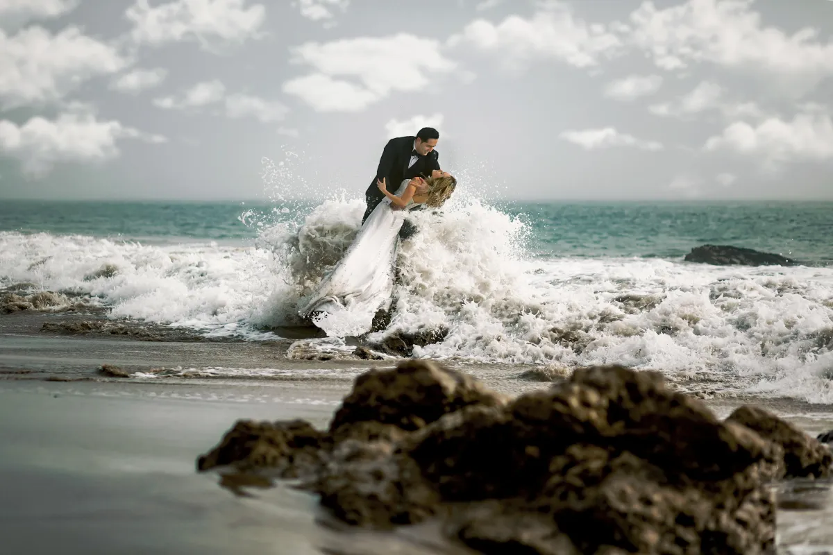 A bride in a flowing white wedding dress and a groom in a tuxedo embracing amidst a large ocean wave breaking on a rocky beach, under a cloudy sky.