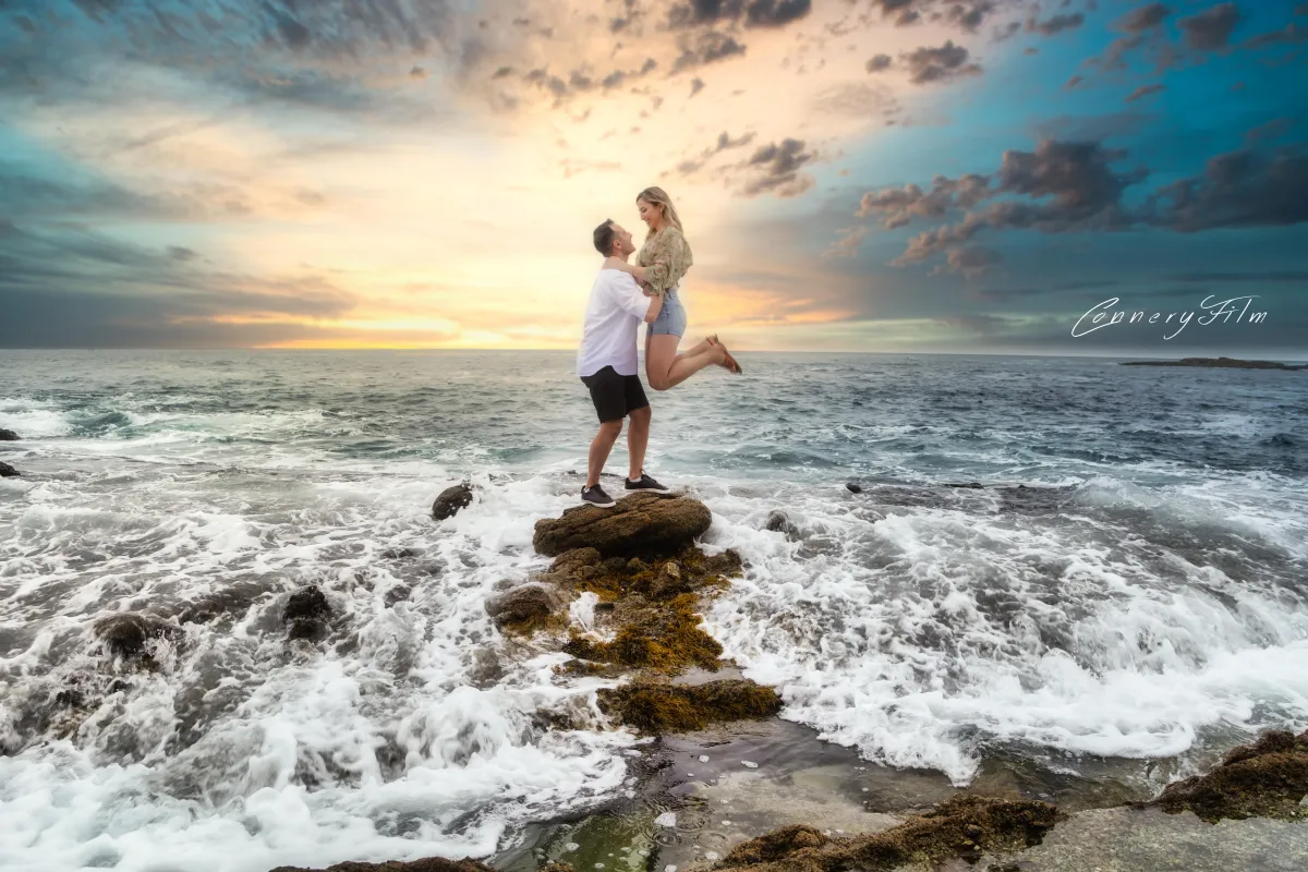 Couple embracing on seaside rocks during sunset, ocean waves crashing around them in a romantic beach setting