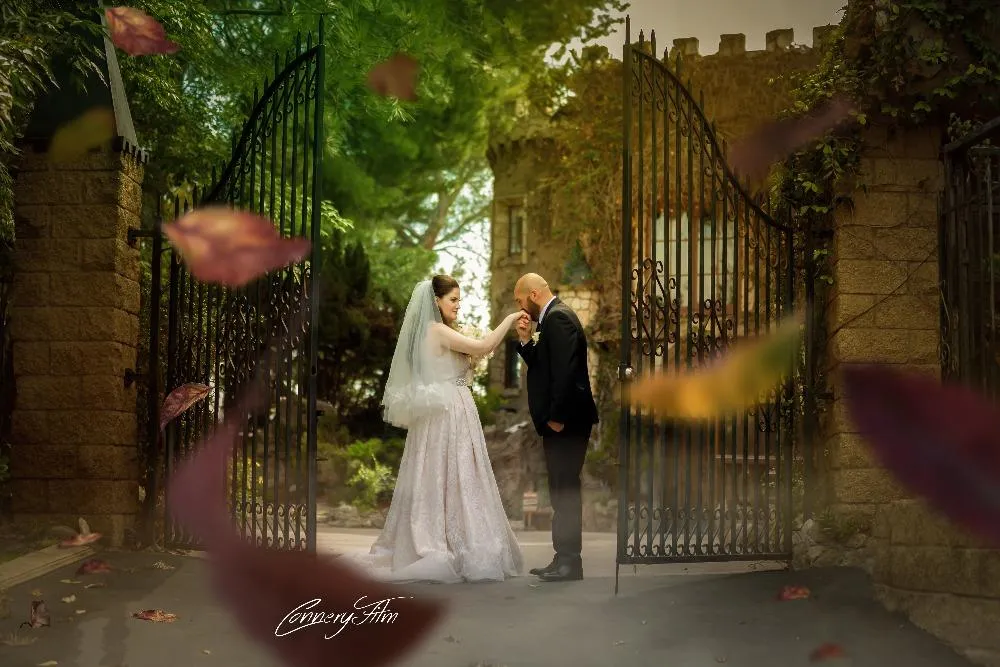 A groom in a tuxedo kissing the hand of his bride in a white wedding gown and veil, standing at the entrance of a grand estate with open wrought iron gates, with autumn leaves falling.