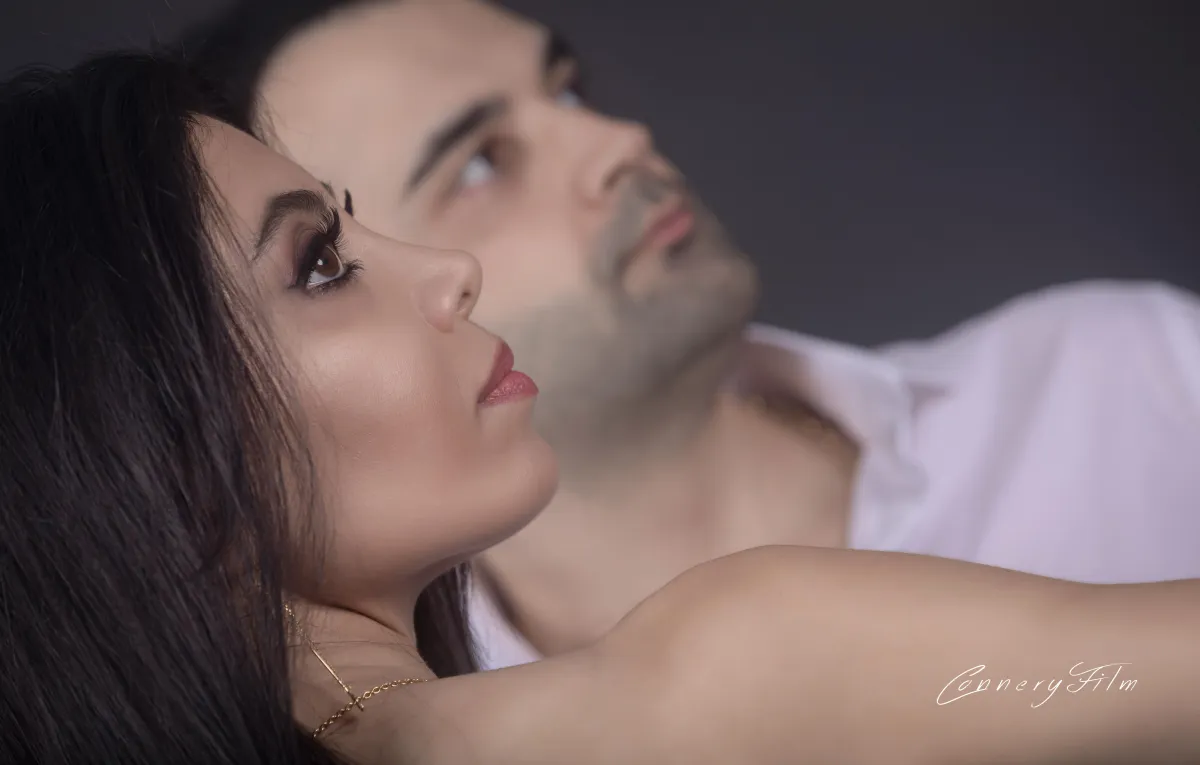 Close-up, artistic portrait of a couple in profile looking upwards, with the woman in focus in the foreground and the man slightly out of focus behind her.