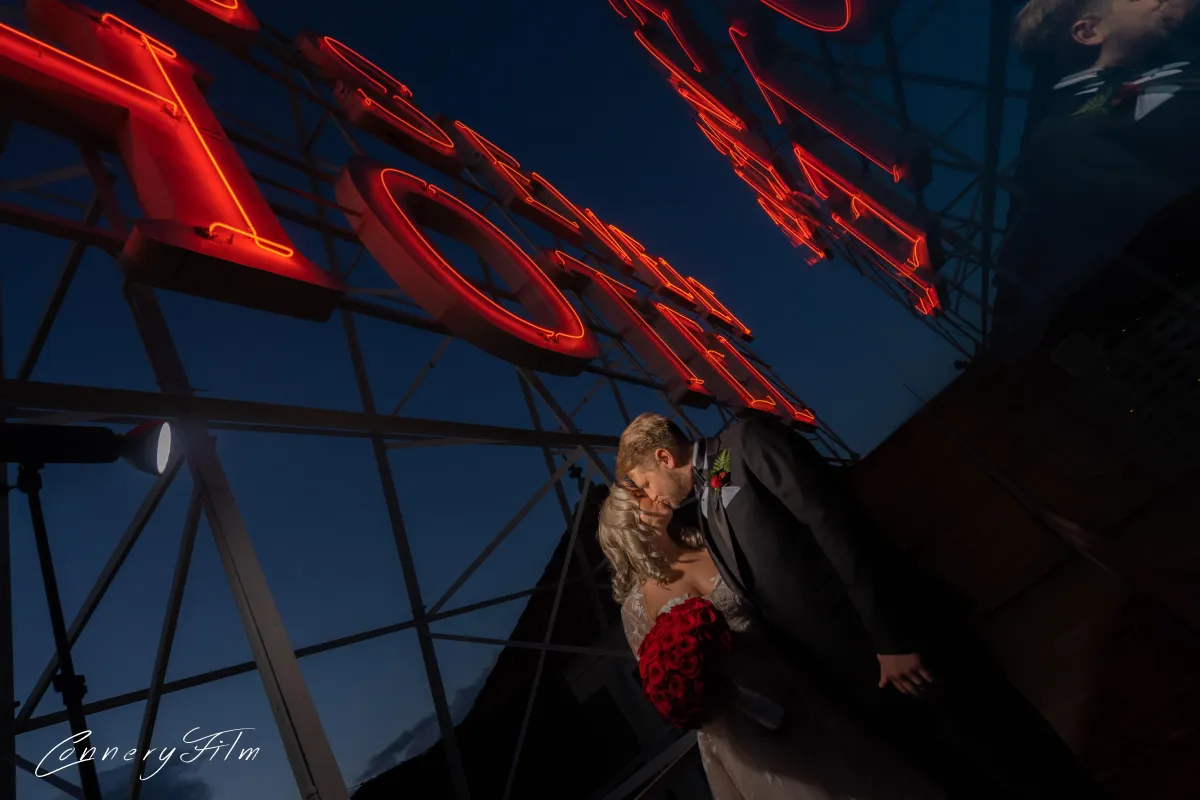 A bride and groom kissing passionately under large red neon "Hollywood" letters at night, with the groom in a tuxedo and the bride holding a red rose bouquet, showcasing a glamorous wedding photo.