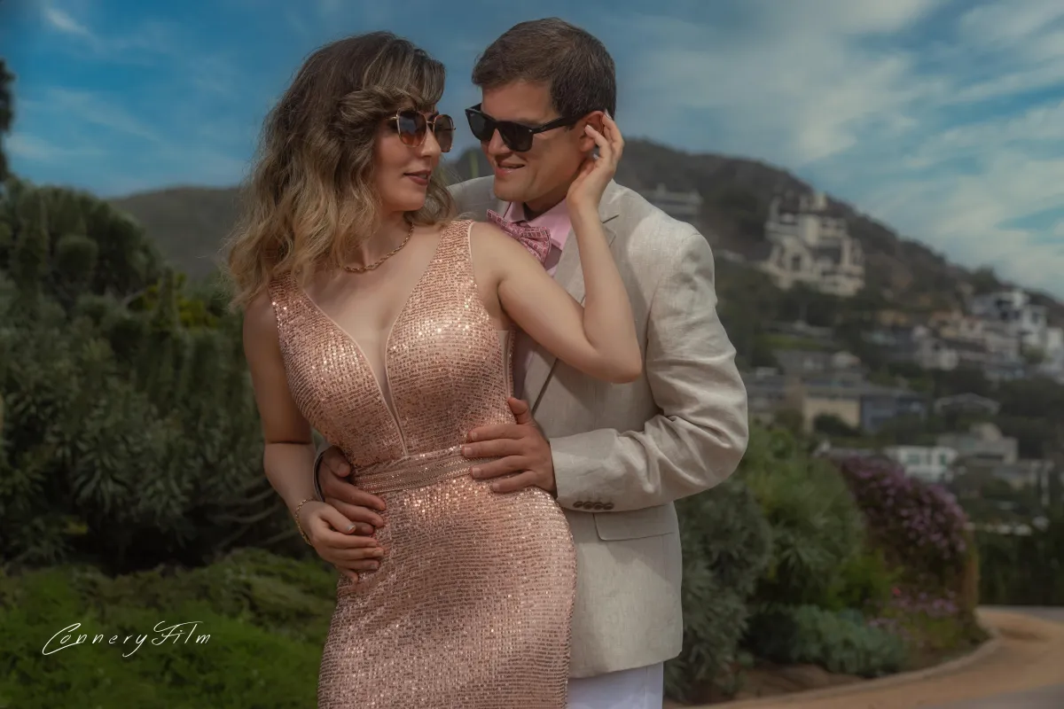 A glamorous couple in stylish attire and sunglasses, embracing outdoors with a scenic background, showcasing their fun and elegant personalities.