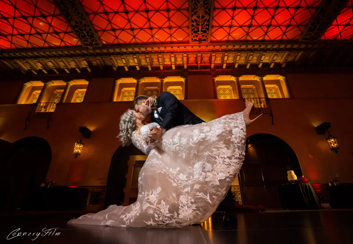 A bride in a long-sleeved lace wedding dress being dipped by her groom in a tuxedo during their first dance, under a beautifully lit, intricate ceiling.