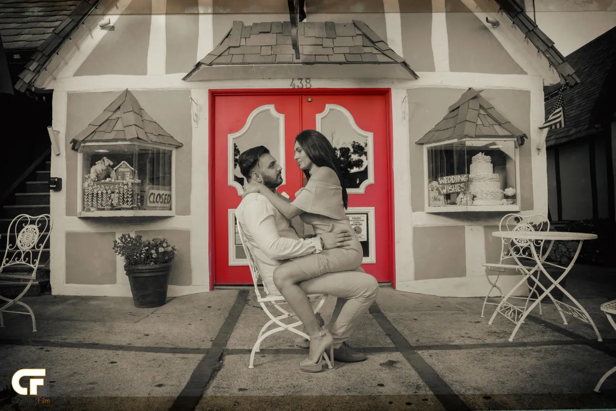 A stylish couple embracing in front of a charming storefront with bright red doors, captured in a black and white photo with selective color on the red elements, conveying romance and fun.