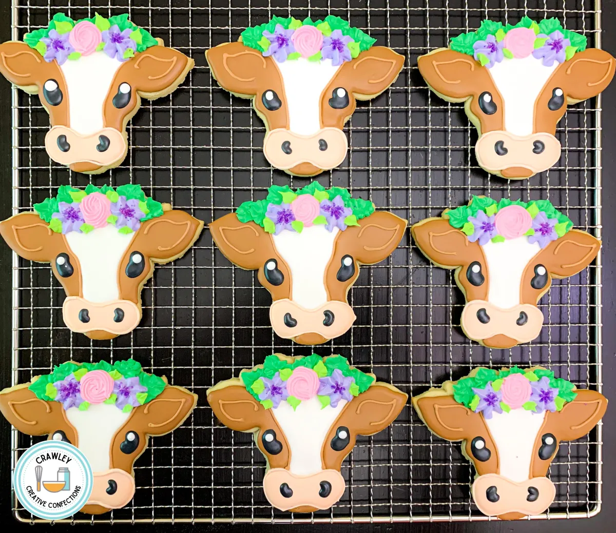 A whimsical collection of sugar cookies shaped like brown and white cows, each decorated with hand-iced details to highlight their distinctive markings. The cows are adorned with vibrant floral crowns in colors like pink, purple, and green, adding a playful and elegant touch. The intricate designs bring these charming farm animals to life in cookie form.