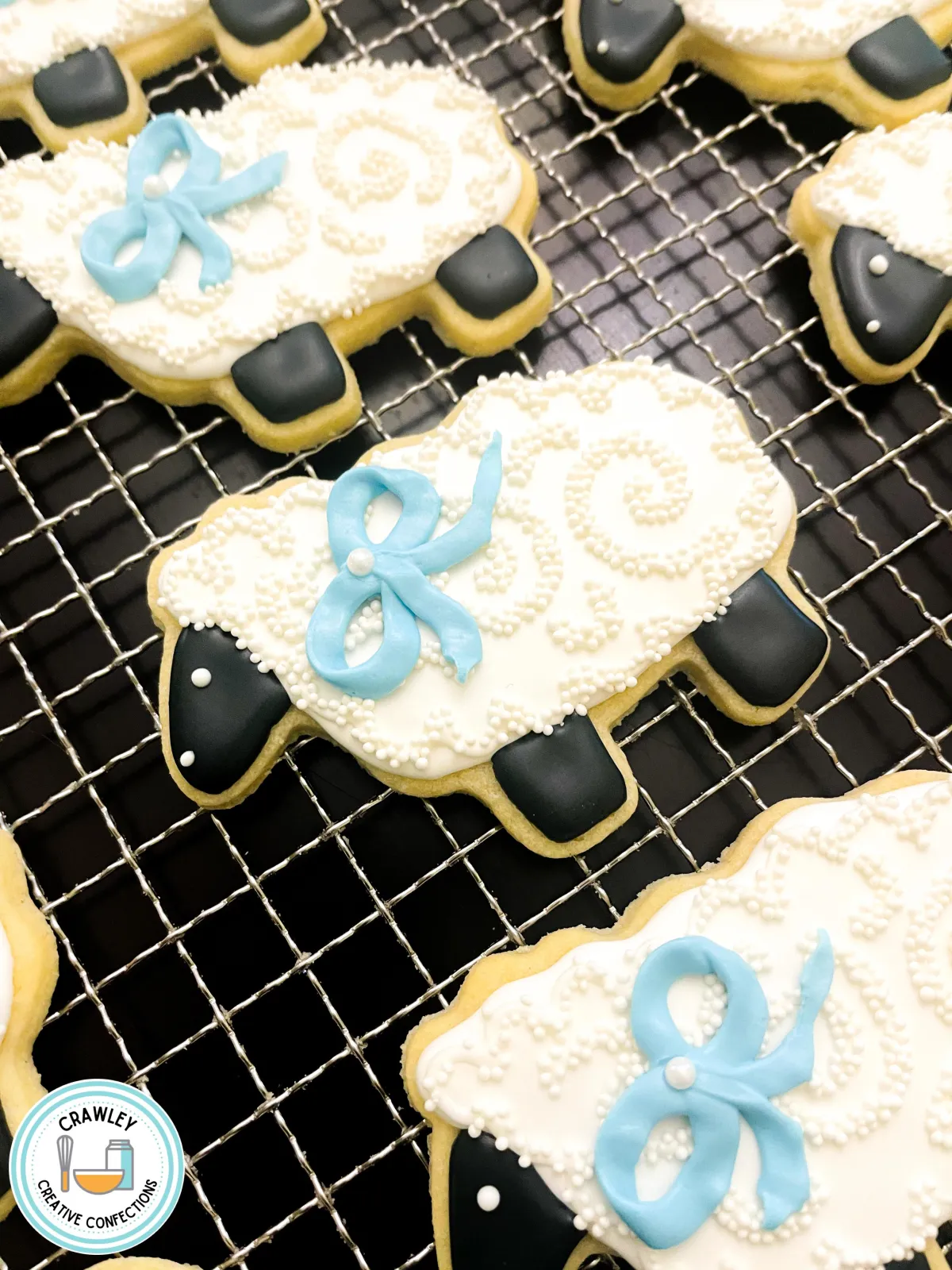 An adorable collection of sugar cookies shaped as fluffy lambs, each decorated with textured white icing to mimic soft wool. The lambs are adorned with delicate blue ribbons around their necks, adding a sweet and charming touch. The hand-iced details bring the whimsical designs to life, perfect for a playful and elegant theme.
