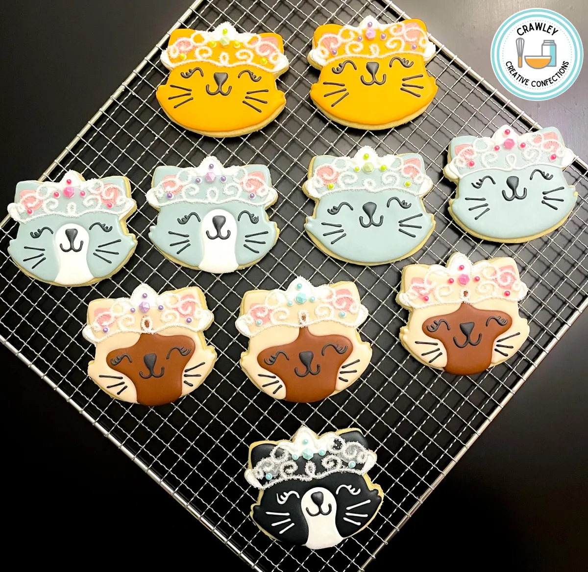 A charming collection of sugar cookies shaped as cat princesses, each decorated to represent different cat breeds. The designs feature elegant tiaras and royal details, paired with unique fur patterns like calico, tabby, Siamese, and solid colors. Each cookie is hand-iced with intricate accents, capturing the personality and beauty of every breed in a whimsical, regal style.