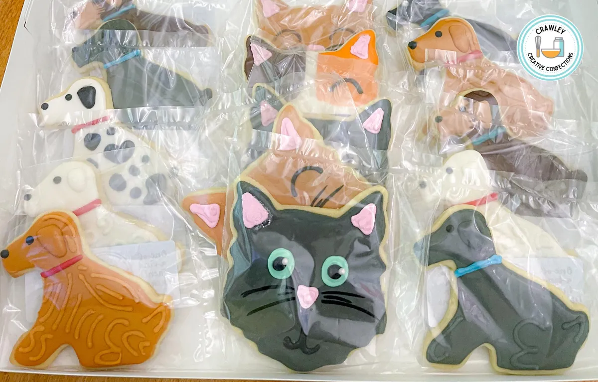 A charming assortment of sugar cookies shaped like cat faces, each decorated to represent different general cat breeds. The designs feature intricate hand-iced details, showcasing tabby stripes, calico patches, Siamese markings, solid colors, and fluffy fur textures. Each cookie captures the unique personality and features of beloved feline companions.