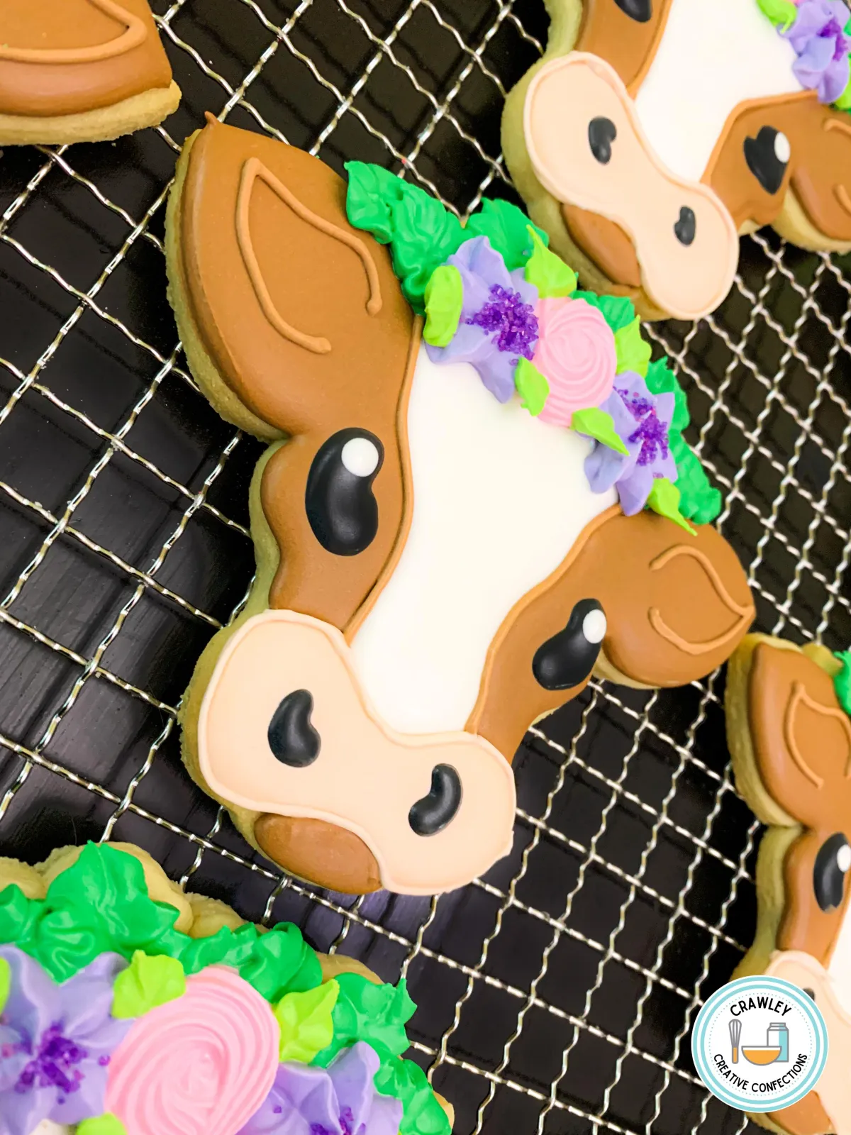 A whimsical collection of sugar cookies shaped like brown and white cows, each decorated with hand-iced details to highlight their distinctive markings. The cows are adorned with vibrant floral crowns in colors like pink, purple, and green, adding a playful and elegant touch. The intricate designs bring these charming farm animals to life in cookie form.