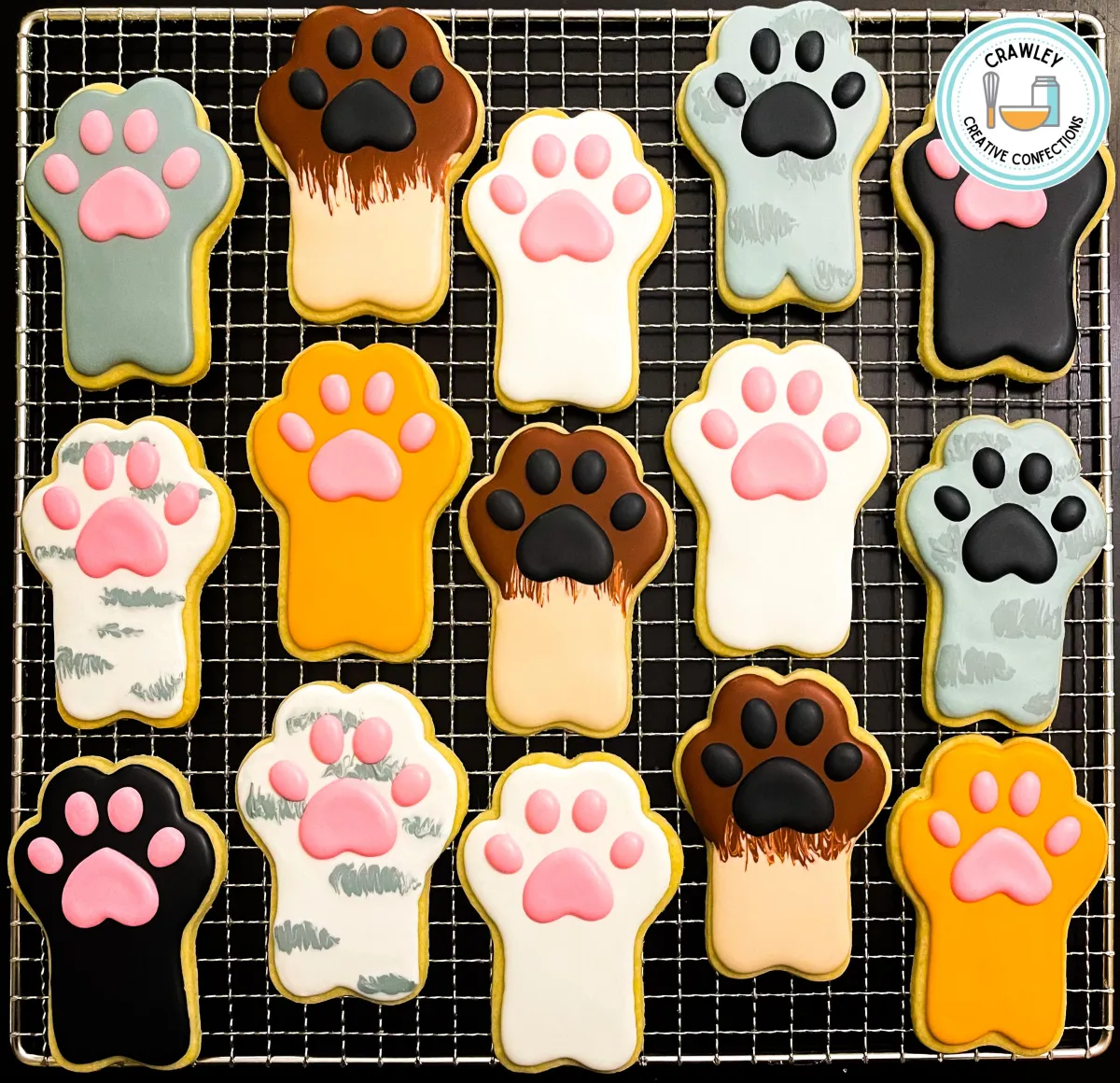 A playful assortment of sugar cookies shaped like cat paws, each decorated to represent different cat breeds. The designs feature a variety of fur patterns and colors, including tabby stripes, calico patches, solid black, and Siamese markings. Each cookie is hand-iced with meticulous detail, creating a charming tribute to feline diversity.