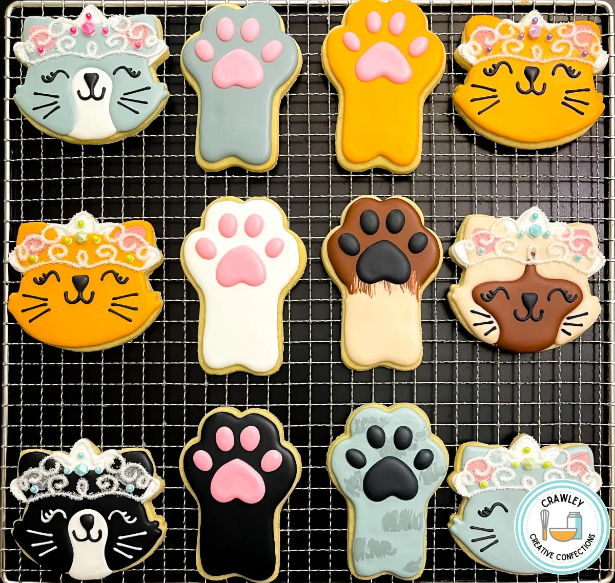 A whimsical collection of cat-themed sugar cookies, featuring elegant princess cats adorned with glittery tiaras and charming details. The set also includes adorable paw print cookies, all beautifully hand-iced in soft pastel colors with intricate accents, creating a purr-fectly royal theme.