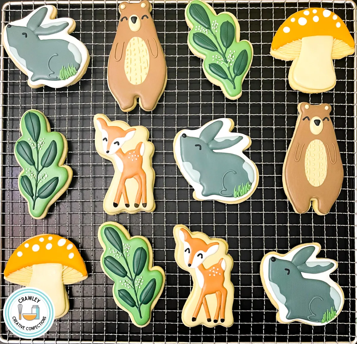 A delightful assortment of woodland animal sugar cookies, featuring intricately hand-iced designs of bears, deer, bunnies, greenery, and mushrooms. Each cookie showcases vibrant colors and detailed textures, surrounded by a rustic backdrop of greenery and pinecones for a cozy, forest-inspired vibe.