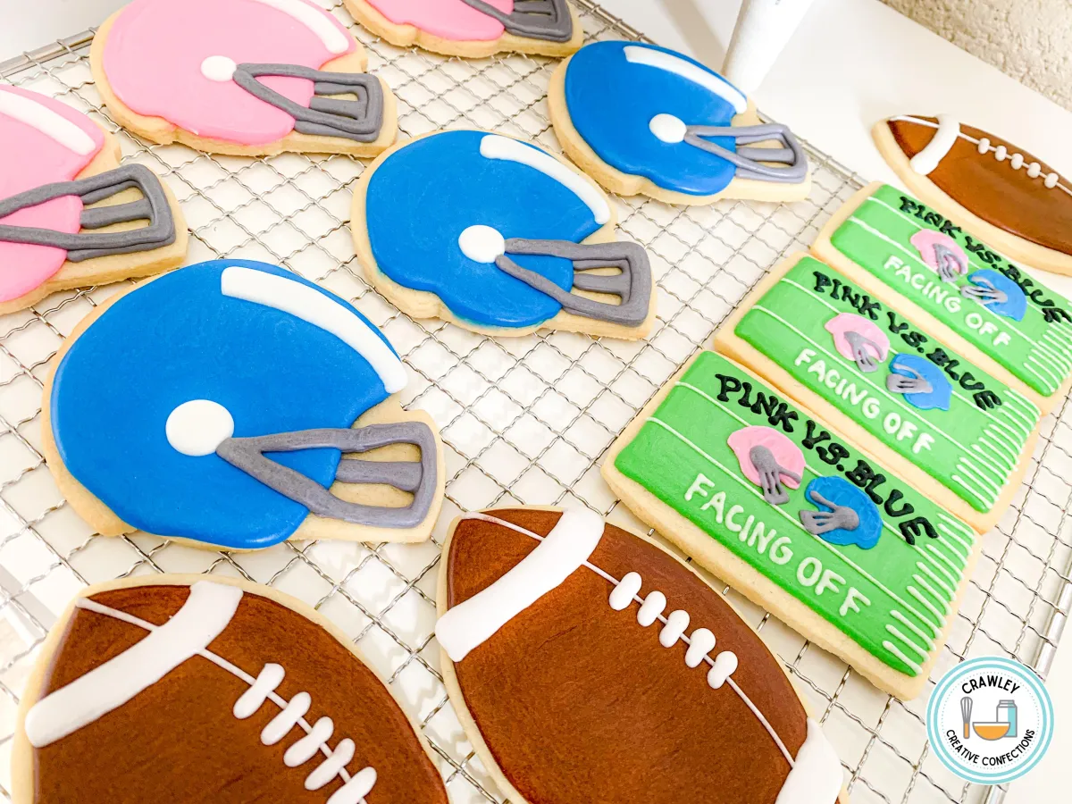 A fun and competitive set of sugar cookies for a gender reveal party featuring a “Pink vs. Blue” theme. The designs include hand-iced football helmets in pink and blue, along with detailed football-shaped cookies. Each cookie captures the playful spirit of the event, blending sports and celebration.