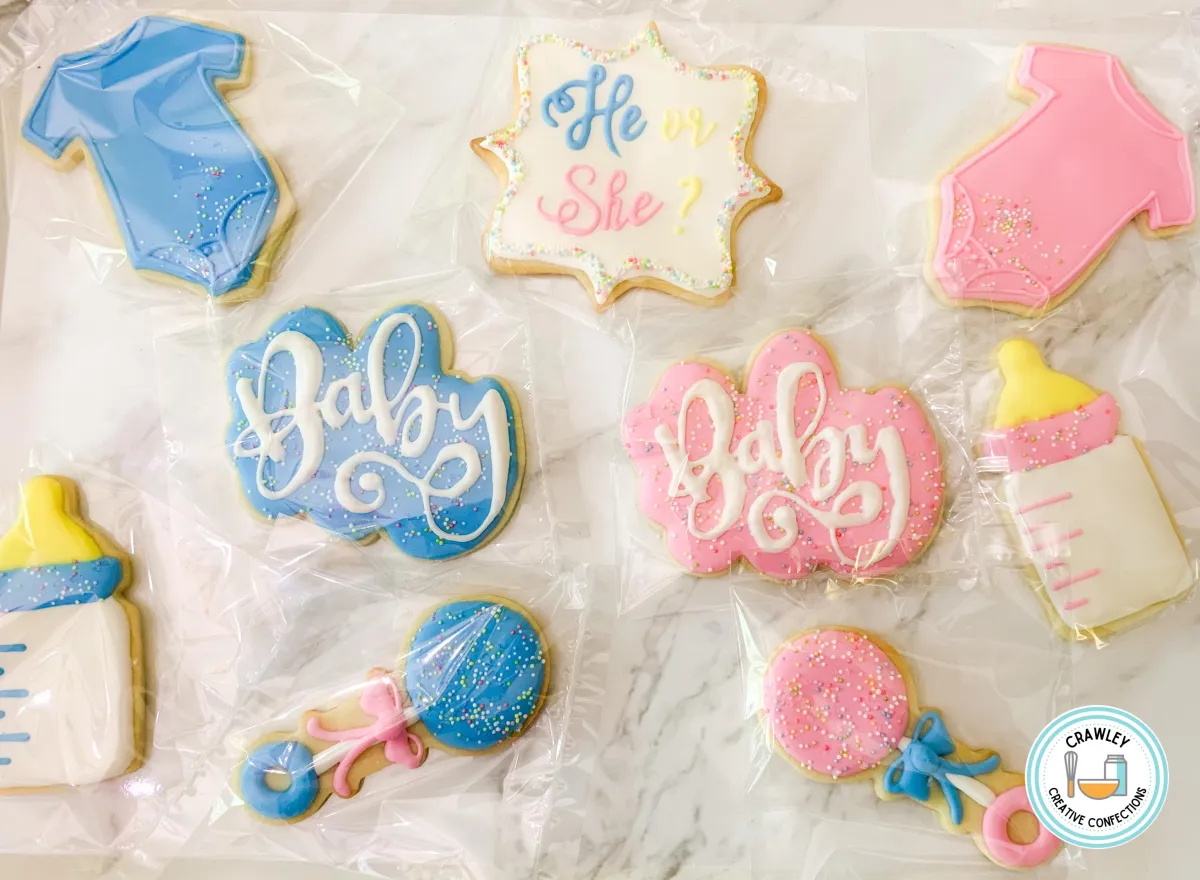 A festive set of sugar cookies for a gender reveal party, decorated with blue, pink, and multi-colored sprinkles. The designs include baby bottles, rattles, and onesies, all hand-iced with intricate details and playful accents, creating a colorful and joyful celebration theme.