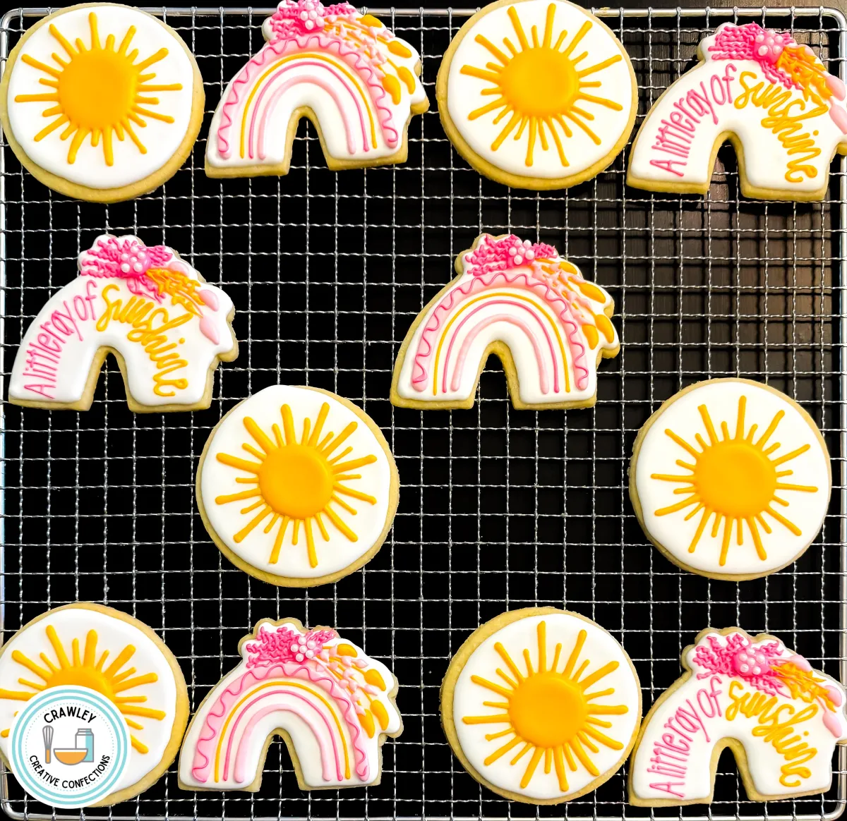 A set of custom sugar cookies displayed on a cooling rack, featuring sunshine and boho rainbow designs in shades of pink, yellow, and white. Some cookies include the phrase “A little ray of sunshine,” with intricate floral accents. These handcrafted cookies by Crawley Creative Confections are perfect for baby showers, birthdays, or cheerful celebrations.