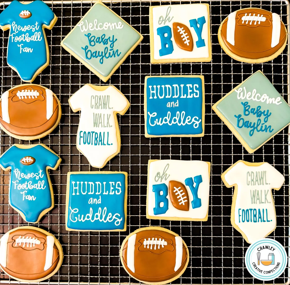 A set of custom football-themed baby shower cookies displayed on a cooling rack. The decorated sugar cookies include footballs, onesies, and square designs with messages such as “Newest Football Fan,” “Oh Boy,” “Huddles and Cuddles,” and “Crawl. Walk. Football.” in shades of blue, white, and green. Made by Crawley Creative Confections, these cookies are perfect for a sports-inspired baby shower or gender reveal.