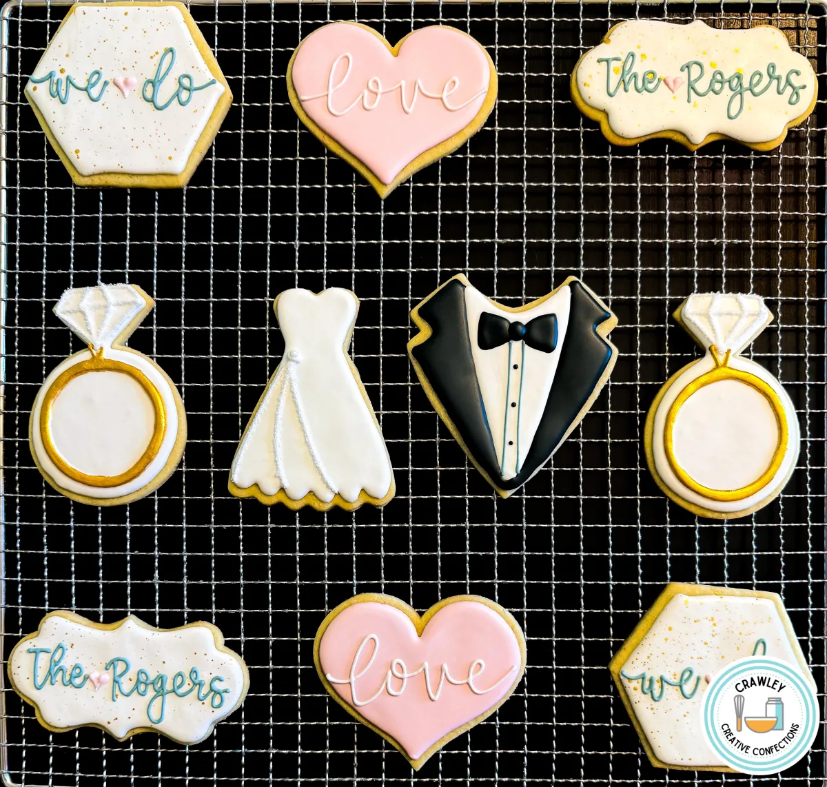 A set of custom wedding-themed sugar cookies displayed on a cooling rack, featuring designs of engagement rings, a wedding dress, a tuxedo, hearts with “love,” and name plaques reading “The Rogers” and “we do.” The cookies are decorated in white, gold, blush pink, and black. Created by Crawley Creative Confections, perfect for weddings, bridal showers, or engagement celebrations.