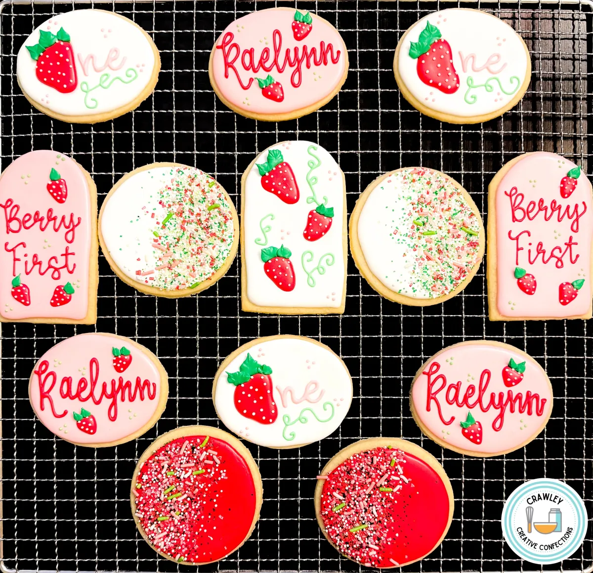 A set of custom strawberry-themed first birthday sugar cookies displayed on a cooling rack. The set includes cookies with hand-piped strawberries, “Berry First” text, “One” designs, personalized “Raelynn” name plaques, and red and white sprinkle-covered cookies. Perfect for a first birthday, baby shower, or summer-themed celebration. Designed by Crawley Creative Confections in Louisville, KY.