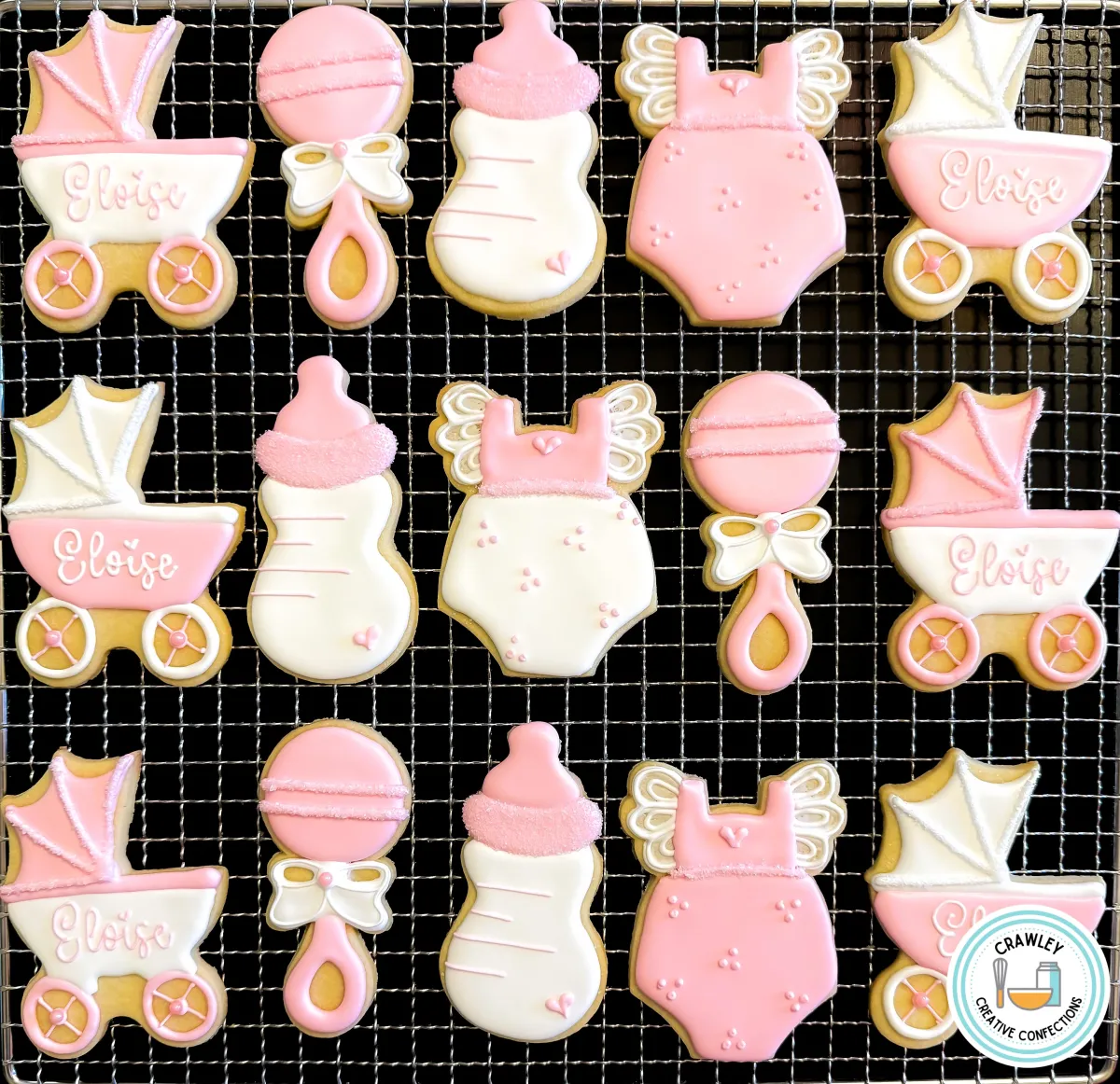 A set of custom pink and gold baby shower sugar cookies displayed on a cooling rack. The collection includes baby carriage cookies personalized with “Eloise,” baby bottles, onesies with angel wings, and rattles, all decorated with delicate details and shimmer accents. Designed for an elegant baby shower, gender reveal, or sip & see party. Created by Crawley Creative Confections in Louisville, KY.