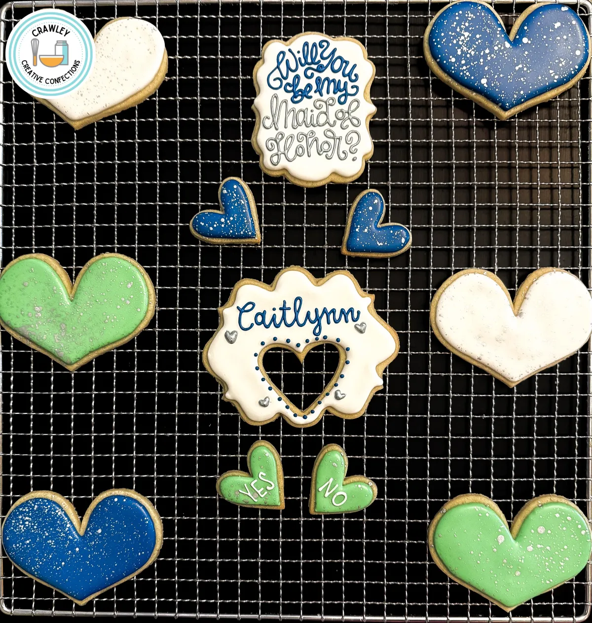 A set of custom bridesmaid proposal sugar cookies displayed on a cooling rack. The collection includes a “Will You Be My Maid of Honor?” cookie, a personalized name plaque with a heart cutout, shimmering blue, green, and white hearts, and a fun “Yes/No” decision cookie. Designed for bridesmaid proposals, engagement parties, or wedding party gifts. Created by Crawley Creative Confections in Louisville, KY.