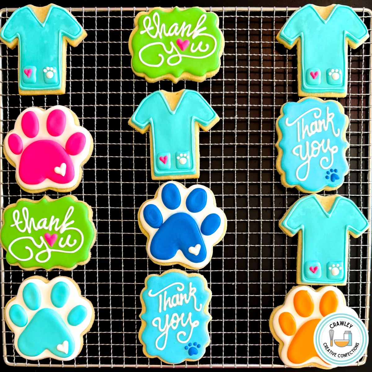 A set of custom veterinary-themed sugar cookies displayed on a cooling rack. The collection includes cookies shaped like scrubs with paw print details, colorful paw print cookies in blue, pink, and orange, and “Thank You” plaques in green and blue. Perfect for vet clinics, animal hospitals, and veterinary appreciation events. Created by Crawley Creative Confections in Louisville, KY.