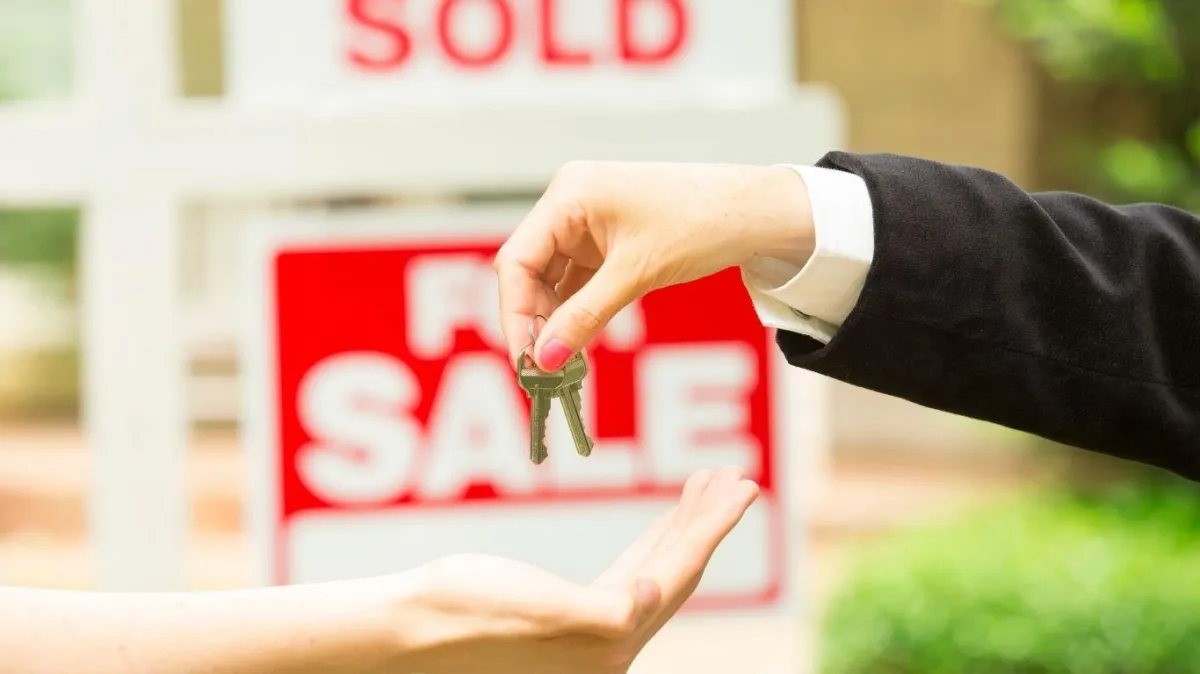 Real estate agent handing house keys to new homeowner in front of a 'For Sale' sign marked 'Sold'