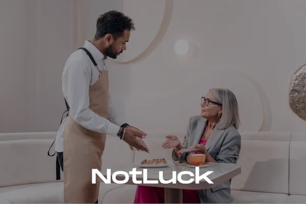 Male waiter in a white shirt and beige apron serves a seated lady with white hair and glasses demonstrating great customer relationship management